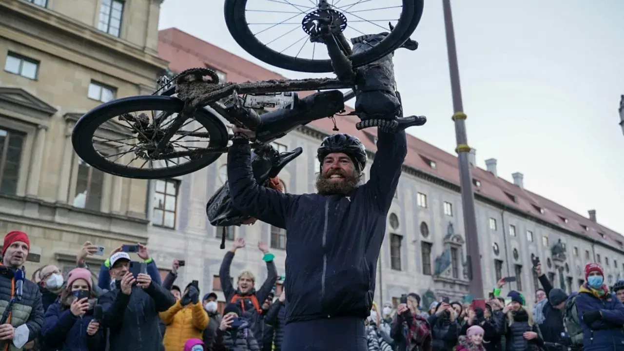 Jonas Deichmann, sichtlich erschöpft, hält sein schlammbedecktes Fahrrad über den Kopf. Eine jubelnde Menge mit Smartphones feiert seine Ankunft.
