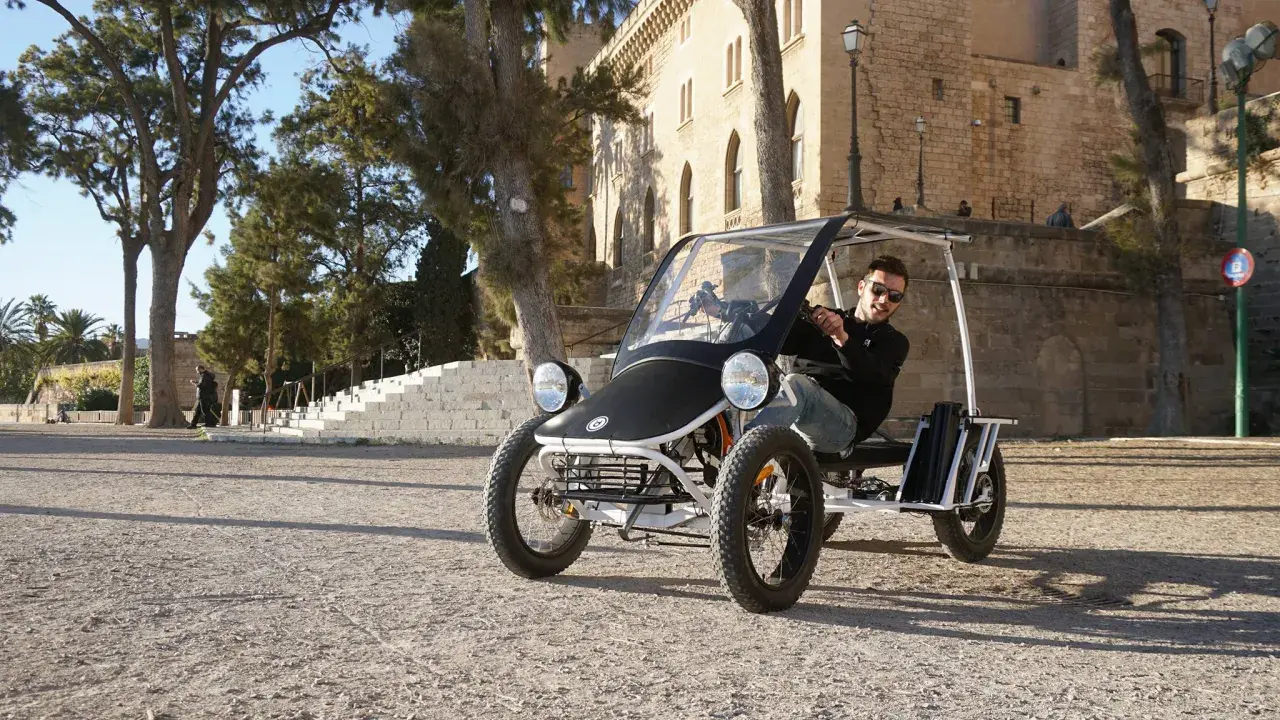 Ein Mann fährt einen Duo Moke durch eine sonnige Gasse mit historischen Gebäuden im Hintergrund.