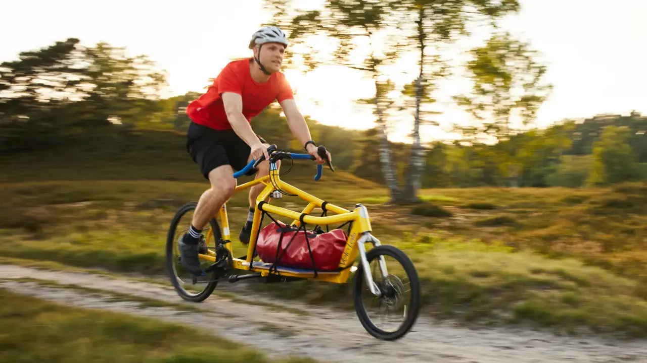 Mann fährt mit einem gelben Velo Lab Lastenrad mit roter Tasche durch eine sonnige Landschaft.