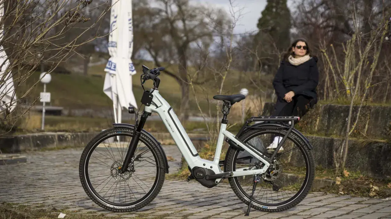 Eine Frau sitzt neben einem hellgrauen Velo de Ville, das auf einem gepflasterten Weg steht. Gute Velo de Ville Erfahrungen sind garantiert.
