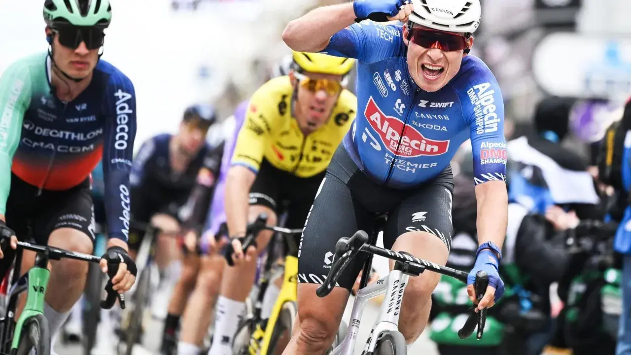 Radfahrer im blauen Trikot von Alpecin-Deceuninck jubelt nach einem Sieg, während andere Fahrer im Peloton folgen.
