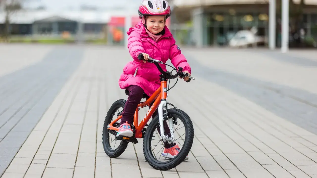 Ein fröhliches Mädchen in pinker Jacke und Helm sitzt auf einem orangefarbenen Kinderfahrrad. Die Frage "kinderfahrrad 4 jahre wieviel zoll" wird durch die Größe des Fahrrads beantwortet.