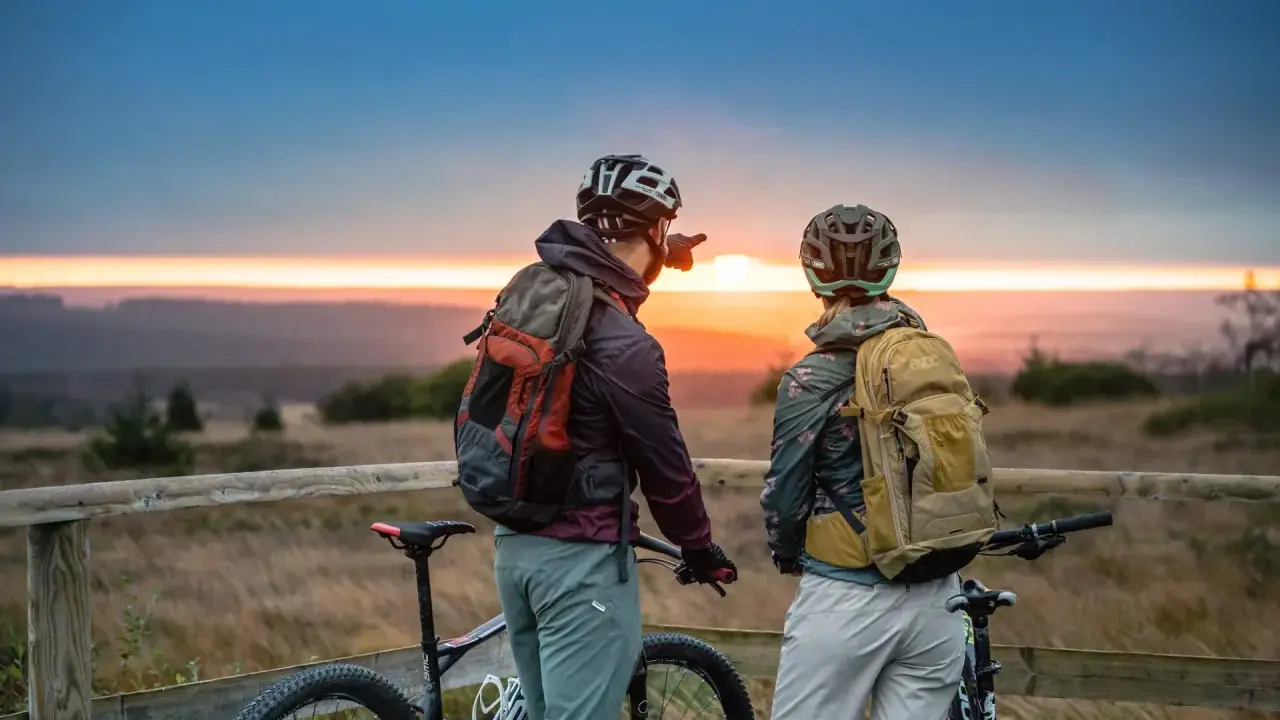 Zwei Radfahrer blicken bei Sonnenuntergang auf die Landschaft. Ein Mann zeigt in die Ferne, vielleicht auf den Stoneman Arduenna.
