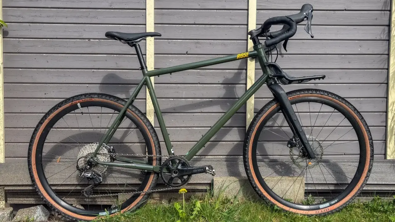 Grünes Rose Hobo Test Gravelbike mit braunen Reifen und Gepäckträger vor Holzwand.