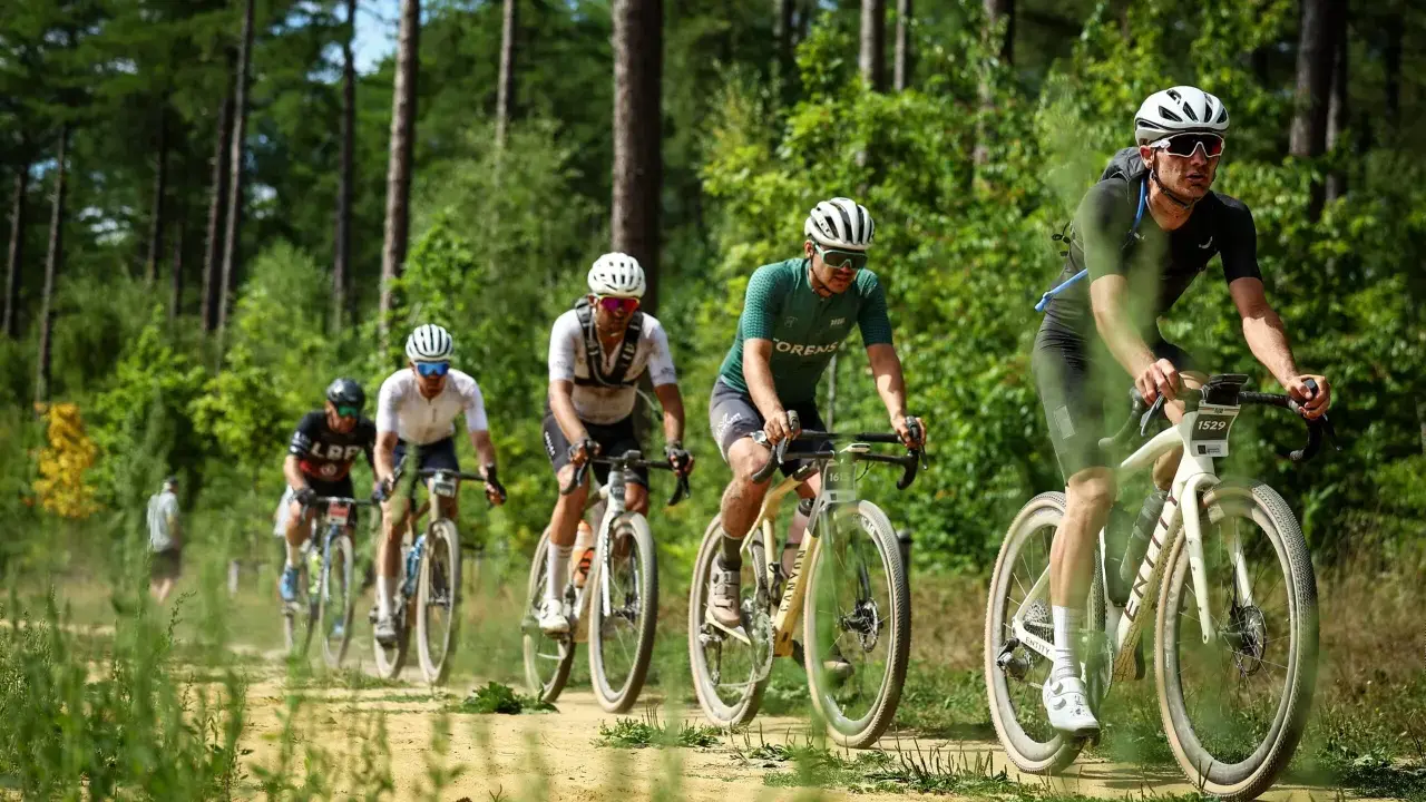 Radfahrer im Wald bei einem Gravel WM-Rennen. Staub wirbelt auf, während sie auf dem sandigen Weg fahren.