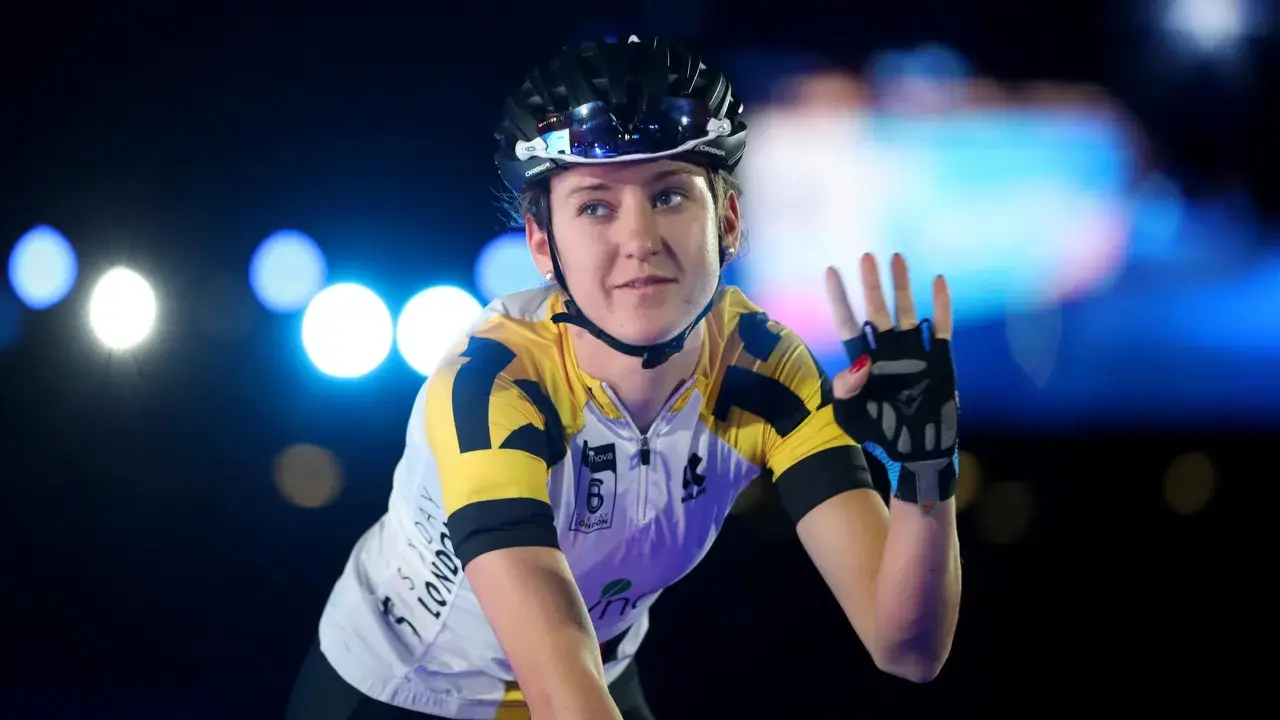 Lea Lin Teutenberg winkt mit der Hand, trägt Radsportkleidung und einen Helm. Blaue und weiße Lichter im Hintergrund.