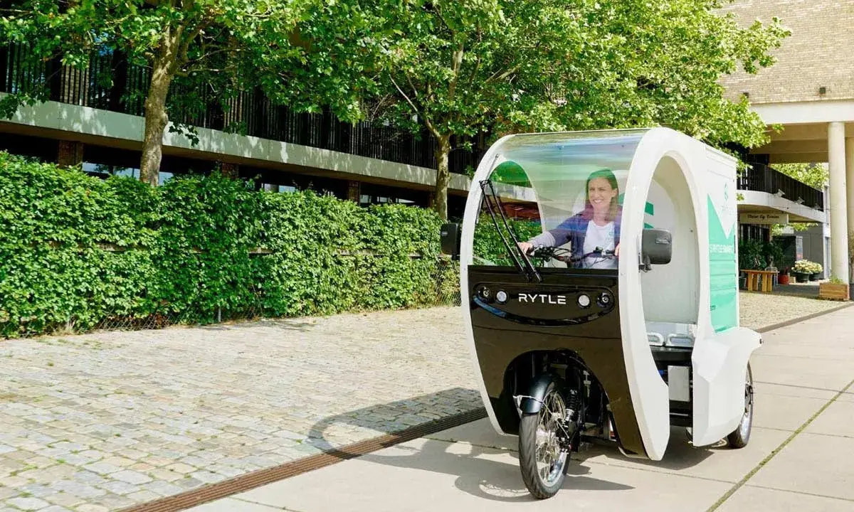 Eine Frau fährt ein Rytle Movr3, ein dreirädriges Elektrofahrzeug, vorbei an einer grünen Hecke und einem Gebäude.