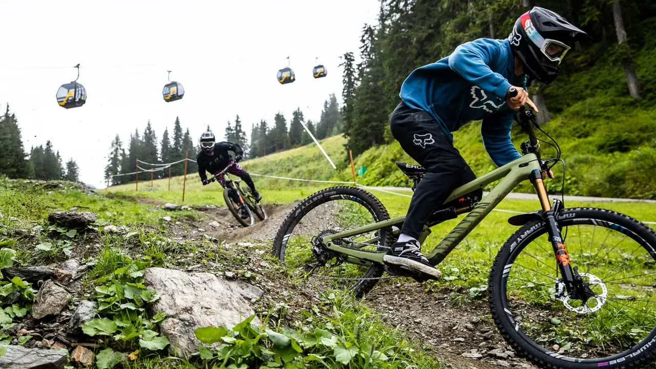 Zwei Mountainbiker auf einem steilen Trail, Seilbahnen im Hintergrund. Einer fährt ein Rad mit Mullet-Setup, was ist ein Mullet?