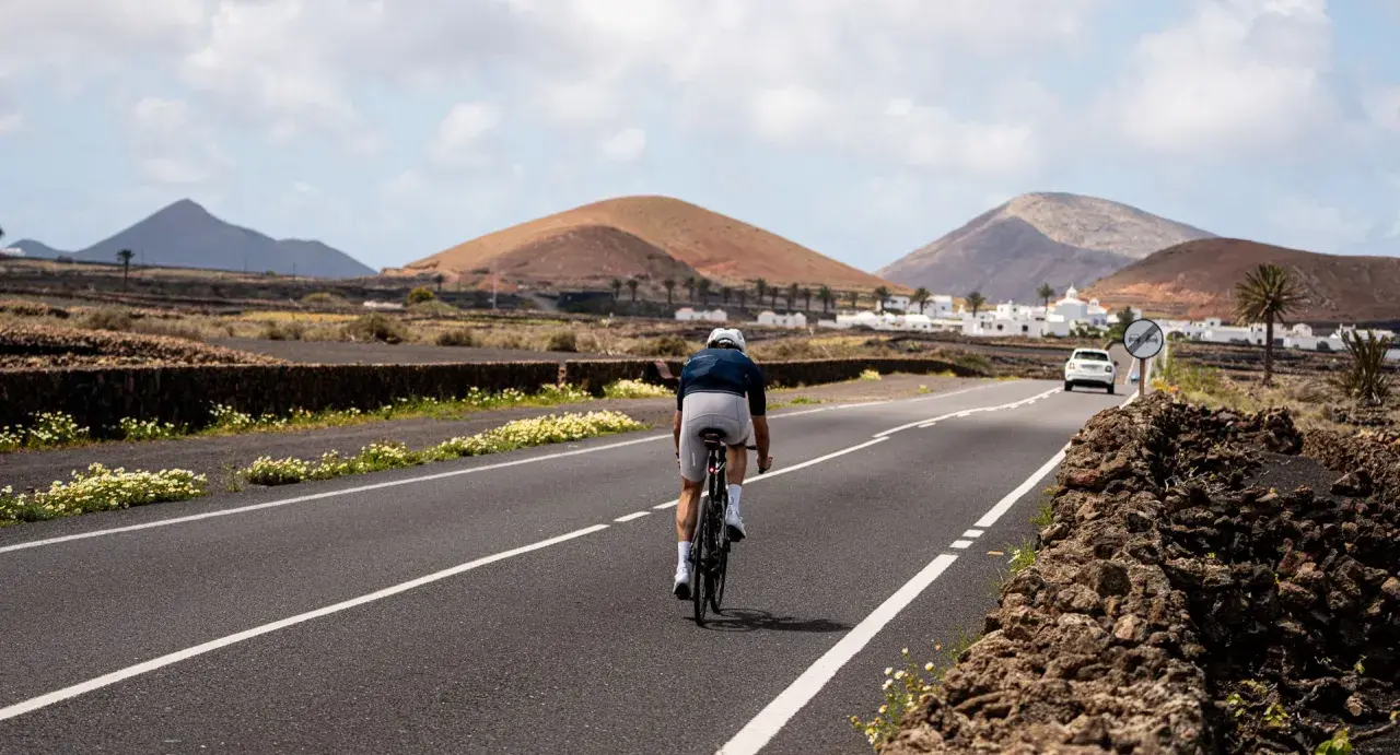Ein Radfahrer auf einem Rennrad fährt auf Lanzarote durch eine vulkanische Landschaft mit weißen Häusern und Palmen.
