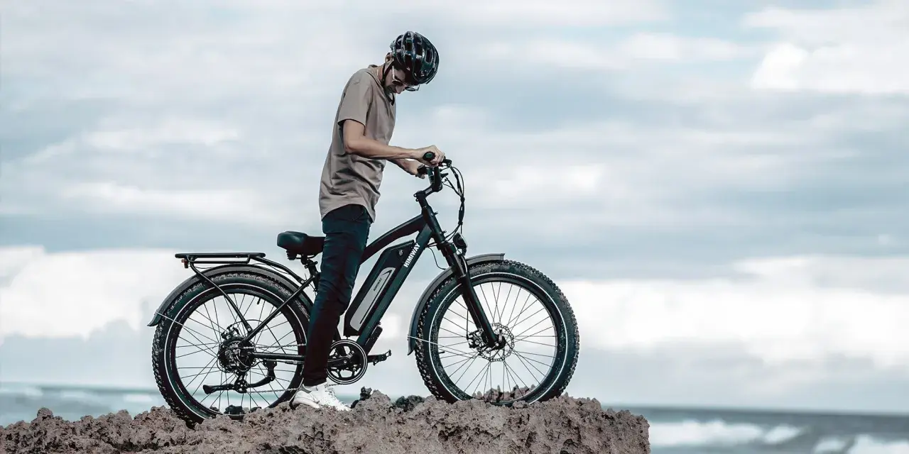 Ein Mann mit Helm steht neben einem Fatbike mit breiten Reifen am Meer. Was ist ein Fatbike? Ein robustes Fahrrad für jedes Gelände.