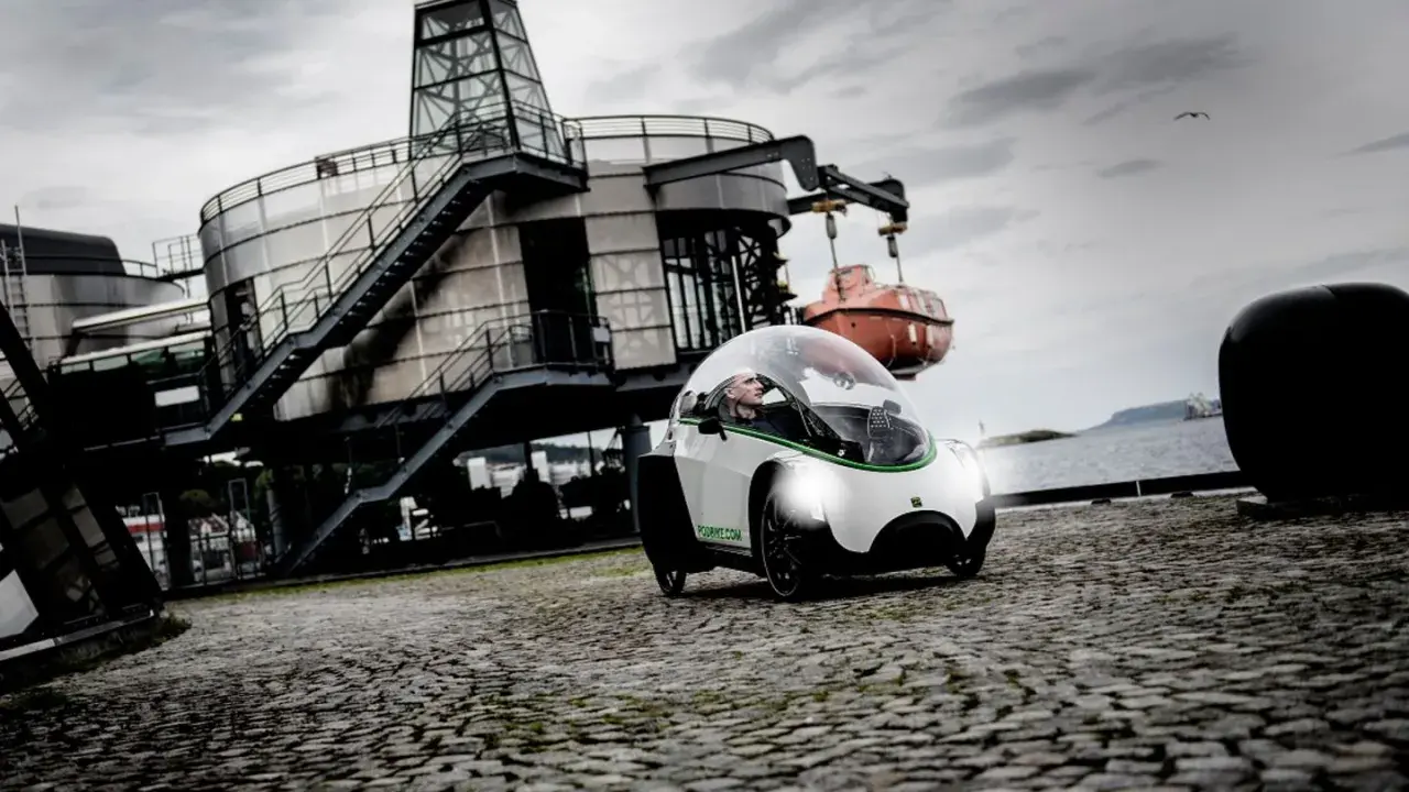 Ein Podbike Frikar fährt auf Kopfsteinpflaster vor einem modernen Gebäude am Wasser.