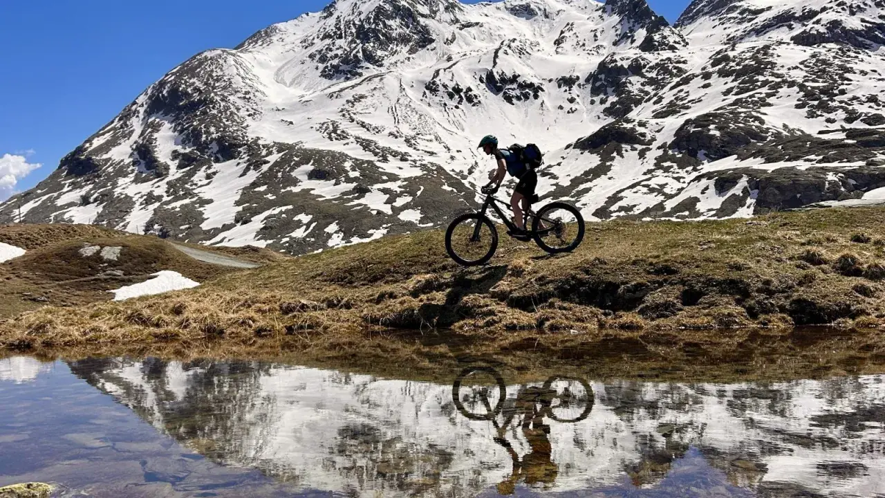 Mountainbiker auf einem epischen Alpencross-Abenteuer, die schneebedeckten Gipfel spiegeln sich in einem Bergsee.