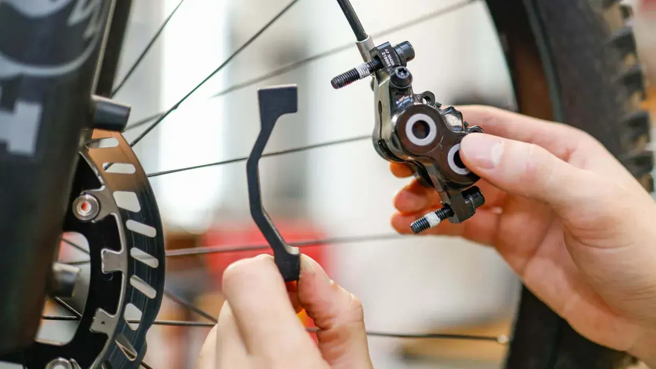 Hände montieren eine Fahrrad-Scheibenbremse. Ein Adapter wird gehalten, um ein Schleifen der Scheibenbremse zu verhindern.