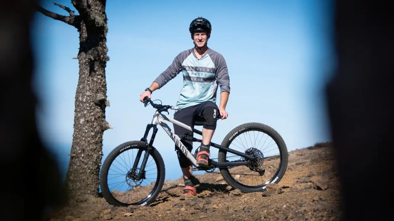 Sam Pilgrim, bereit für die nächste Abfahrt, posiert mit seinem Mountainbike auf einem felsigen Hang vor blauem Himmel.