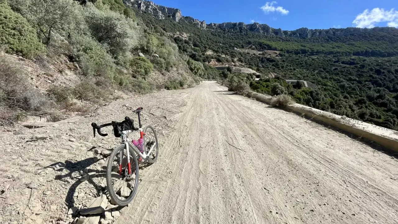 Ein Rennrad steht auf einer staubigen Straße in Sardinien, umgeben von felsigen Hängen und grüner Vegetation.