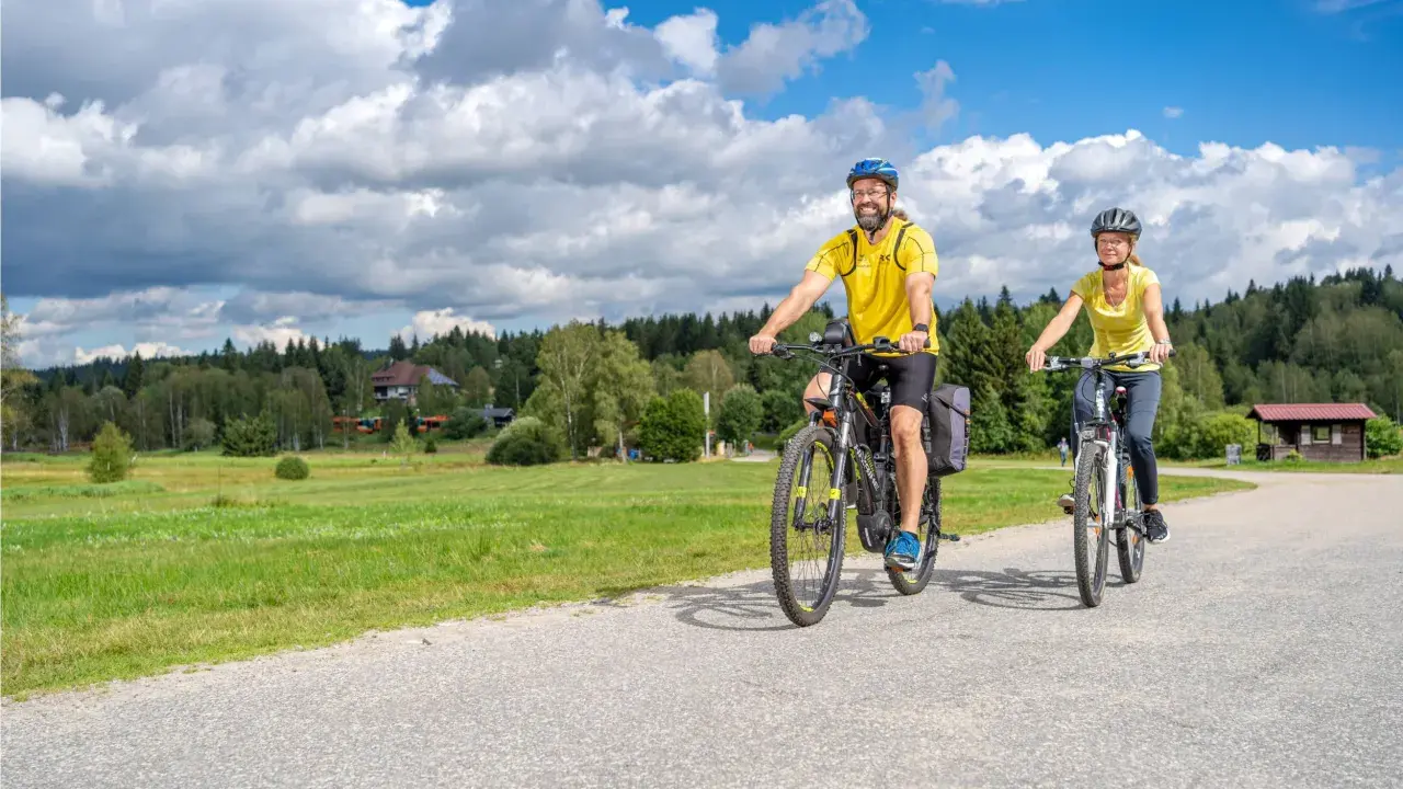 Glückliches Paar auf E-Bikes auf einer Tour durch den Bayerischen Wald. Die Sonne scheint, der Himmel ist blau mit weißen Wolken.