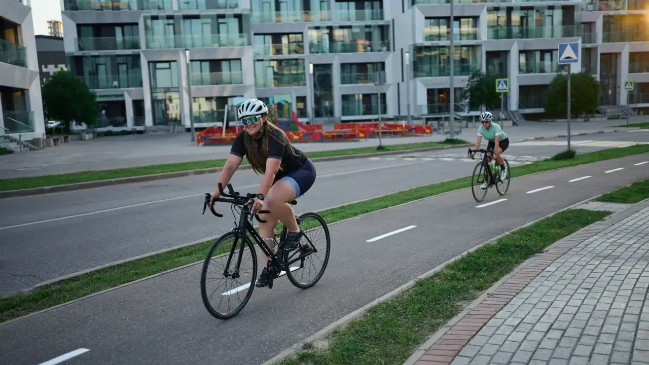 Zwei Rennradfahrerinnen genießen eine Fahrt auf einem Radweg vor modernen Gebäuden.