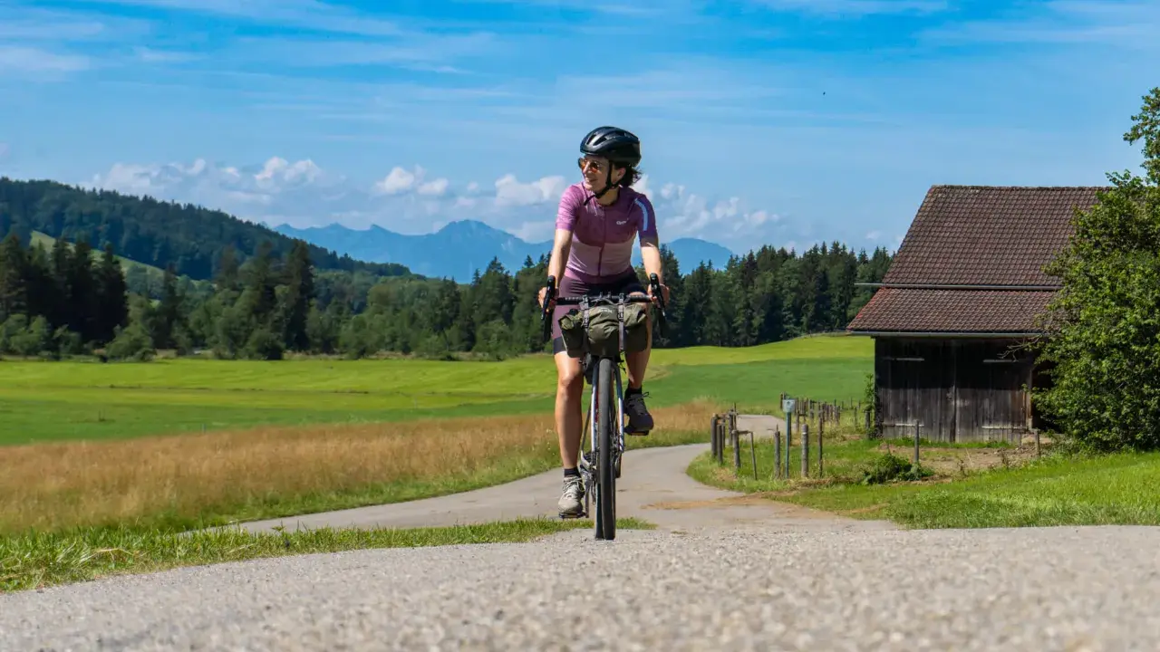 Frau auf einer e bike tour durch grüne Wiesen und Berge, mit einem alten Holzhaus am Wegesrand.