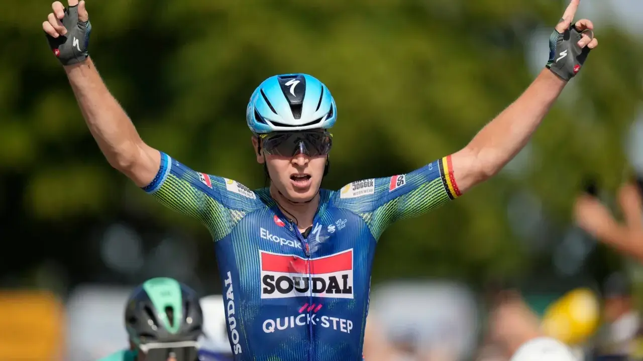 Tim Merlier jubelt mit erhobenen Armen und zeigt den Sieg. Er trägt ein blau-grünes Radtrikot mit Sponsorenlogos.
