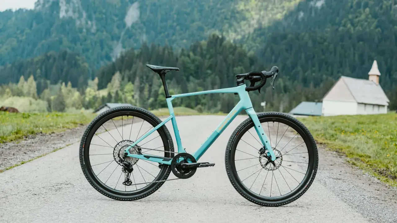 Ein hellblaues Simplon Grid Gravelbike steht auf einer asphaltierten Straße vor einer Bergkulisse mit Wäldern und einer kleinen Kirche.