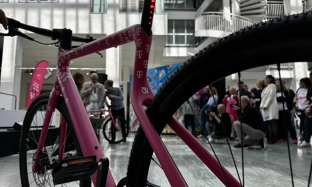 Ein pinkes company bike der Telekom mit vielen T-Logos. Im Hintergrund stehen Menschen und eine Wendeltreppe.