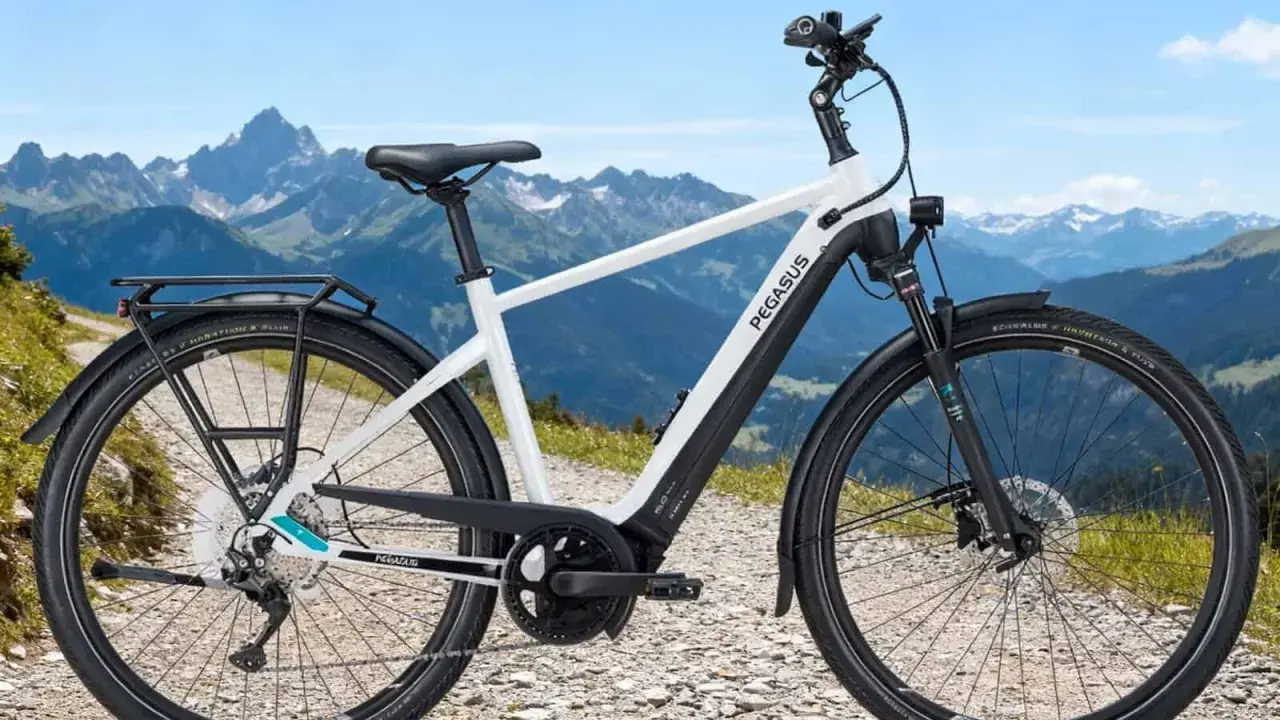 Ein weißes Pegasus E-Bike mit Gepäckträger steht auf einem Schotterweg vor einer Bergkulisse. Perfekt, um das E-Bike zu testen.
