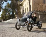 Ein Mann fährt einen Duo Moke durch eine sonnige Gasse mit historischen Gebäuden im Hintergrund.
