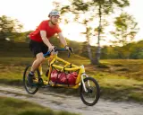 Mann fährt mit einem gelben Velo Lab Lastenrad mit roter Tasche durch eine sonnige Landschaft.