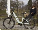 Eine Frau sitzt neben einem hellgrauen Velo de Ville, das auf einem gepflasterten Weg steht. Gute Velo de Ville Erfahrungen sind garantiert.