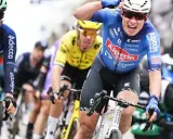 Radfahrer im blauen Trikot von Alpecin-Deceuninck jubelt nach einem Sieg, während andere Fahrer im Peloton folgen.