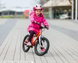 Ein fröhliches Mädchen in pinker Jacke und Helm sitzt auf einem orangefarbenen Kinderfahrrad. Die Frage "kinderfahrrad 4 jahre wieviel zoll" wird durch die Größe des Fahrrads beantwortet.