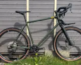 Grünes Rose Hobo Test Gravelbike mit braunen Reifen und Gepäckträger vor Holzwand.