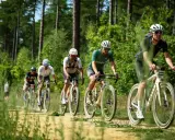 Radfahrer im Wald bei einem Gravel WM-Rennen. Staub wirbelt auf, während sie auf dem sandigen Weg fahren.