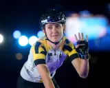 Lea Lin Teutenberg winkt mit der Hand, trägt Radsportkleidung und einen Helm. Blaue und weiße Lichter im Hintergrund.