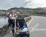 Frau mit Rucksack und Kinderwagen auf einer Brücke, bereit für eine abc tour. Im Hintergrund eine Stadt und Berge.