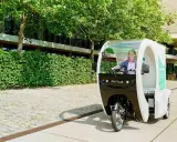 Eine Frau fährt ein Rytle Movr3, ein dreirädriges Elektrofahrzeug, vorbei an einer grünen Hecke und einem Gebäude.