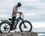 Ein Mann mit Helm steht neben einem Fatbike mit breiten Reifen am Meer. Was ist ein Fatbike? Ein robustes Fahrrad für jedes Gelände.