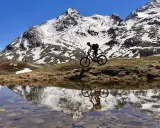 Mountainbiker auf einem epischen Alpencross-Abenteuer, die schneebedeckten Gipfel spiegeln sich in einem Bergsee.