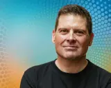 Porträt eines Mannes mit freundlichem Lächeln vor einem bunten Hintergrund. Man fragt sich, was macht Jan Ullrich heute?