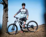 Sam Pilgrim, bereit für die nächste Abfahrt, posiert mit seinem Mountainbike auf einem felsigen Hang vor blauem Himmel.
