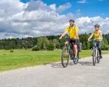 Glückliches Paar auf E-Bikes auf einer Tour durch den Bayerischen Wald. Die Sonne scheint, der Himmel ist blau mit weißen Wolken.