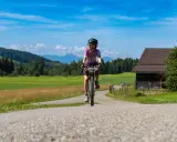 Frau auf einer e bike tour durch grüne Wiesen und Berge, mit einem alten Holzhaus am Wegesrand.