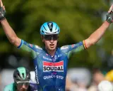 Tim Merlier jubelt mit erhobenen Armen und zeigt den Sieg. Er trägt ein blau-grünes Radtrikot mit Sponsorenlogos.