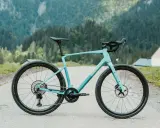 Ein hellblaues Simplon Grid Gravelbike steht auf einer asphaltierten Straße vor einer Bergkulisse mit Wäldern und einer kleinen Kirche.