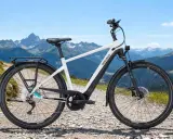 Ein weißes Pegasus E-Bike mit Gepäckträger steht auf einem Schotterweg vor einer Bergkulisse. Perfekt, um das E-Bike zu testen.