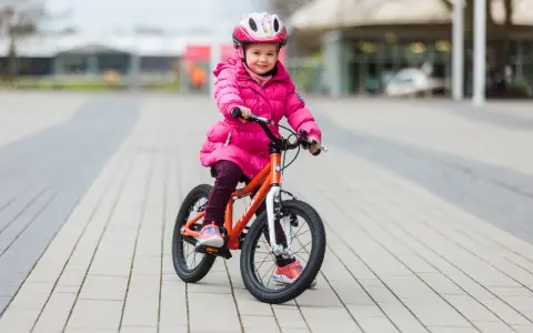 Ein fröhliches Mädchen in pinker Jacke und Helm sitzt auf einem orangefarbenen Kinderfahrrad. Die Frage "kinderfahrrad 4 jahre wieviel zoll" wird durch die Größe des Fahrrads beantwortet.