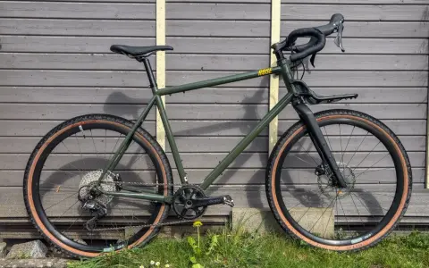 Grünes Rose Hobo Test Gravelbike mit braunen Reifen und Gepäckträger vor Holzwand.