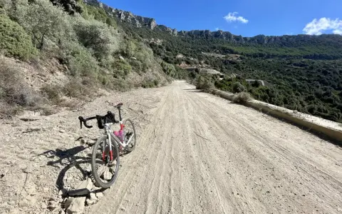 Ein Rennrad steht auf einer staubigen Straße in Sardinien, umgeben von felsigen Hängen und grüner Vegetation.