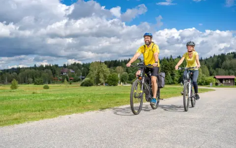 Glückliches Paar auf E-Bikes auf einer Tour durch den Bayerischen Wald. Die Sonne scheint, der Himmel ist blau mit weißen Wolken.