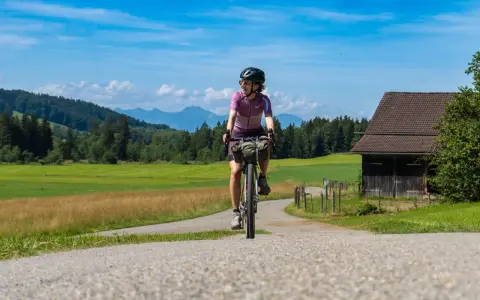 Frau auf einer e bike tour durch grüne Wiesen und Berge, mit einem alten Holzhaus am Wegesrand.
